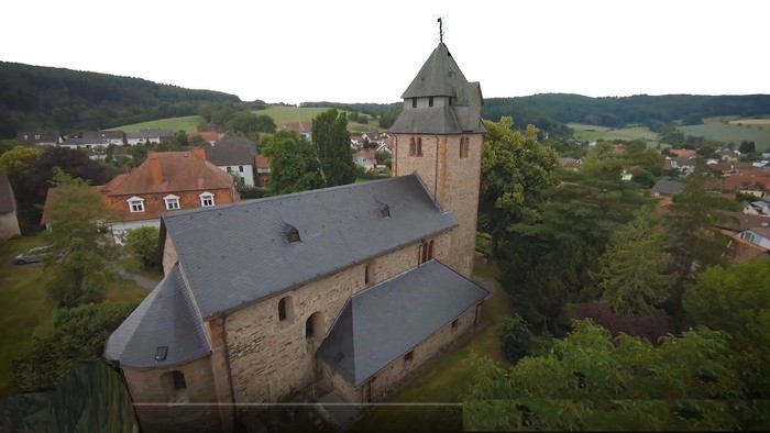 Luftaufnahme einer historischen Kirche mit hohem Turm, umgeben von einem malerischen Dorf und üppigem Grün.