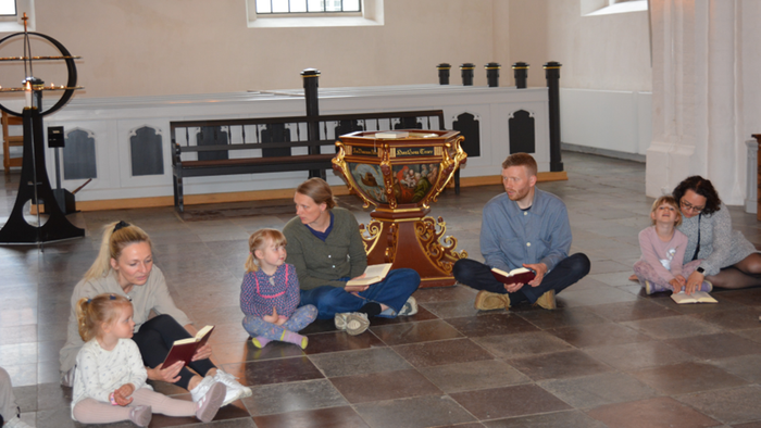 En gruppe af voksne og børn sidder på gulvet i en historisk sal og læser fra bøger.