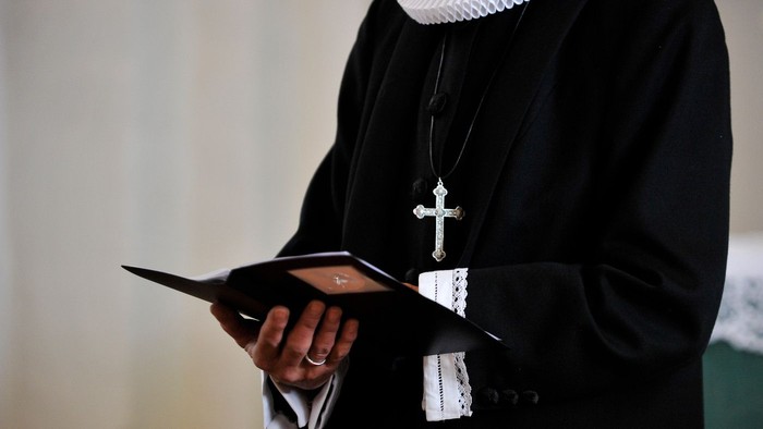 En præst i sort liturgisk klæde holder en bog og en kæde med kors.