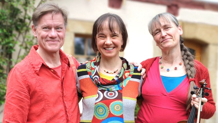 Drei Menschen posieren vor einem Haus, einer hält eine Geige