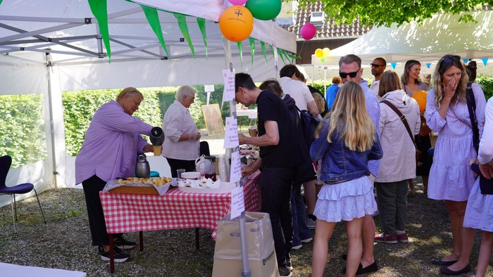 Udendørs sammenkomst med folk, der serverer og nyder mad under et telt med farverige balloner.