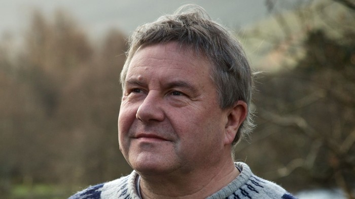 Middle-aged man in sweater standing outdoors near water.