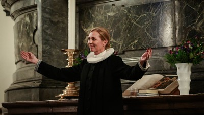 Kvinde i mørkt kapel, der holder forgyldt objekt og har arme i vejr.