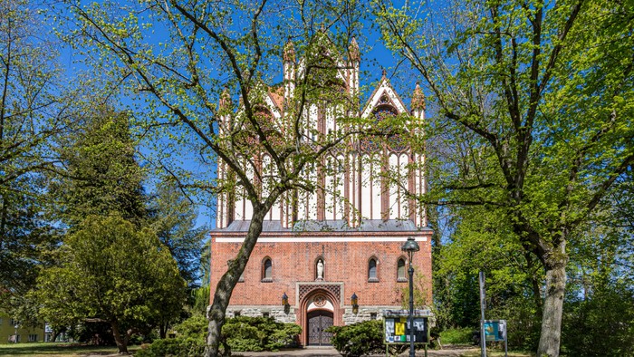 Das Bild zeigt eine alte Kirche mit hohem Turm, umgeben von Bäumen und einem klaren blauen Himmel.