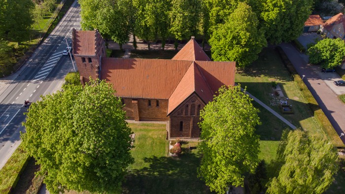 En rødtaget kirke omgivet af grønne træer og en parkering.