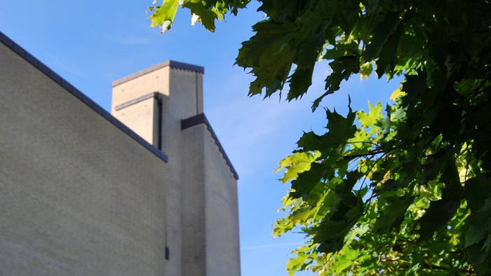 Et højt, kvadratisk tårn med en smal, rektangulær vindue og en kamin, omgivet af grønne blade.