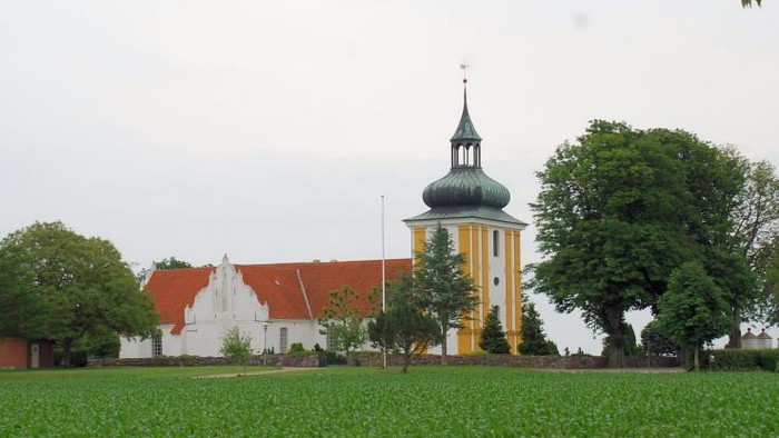 En grøn og åben mark med et gult tårn og røde og hvide bygninger i baggrunden