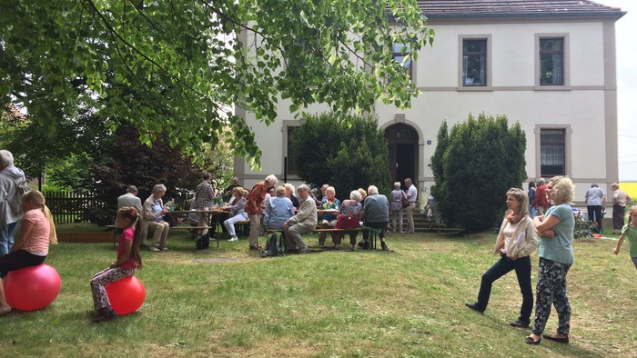 Im Garten vor einem Haus feiern Menschen bei einem Picknick, während Kinder mit bunten Bällen spielen.