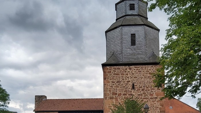 Historische Steinkirche mit modernem Glasanbau, umgeben von Grünflächen und vor bewölktem Himmel gelegen.