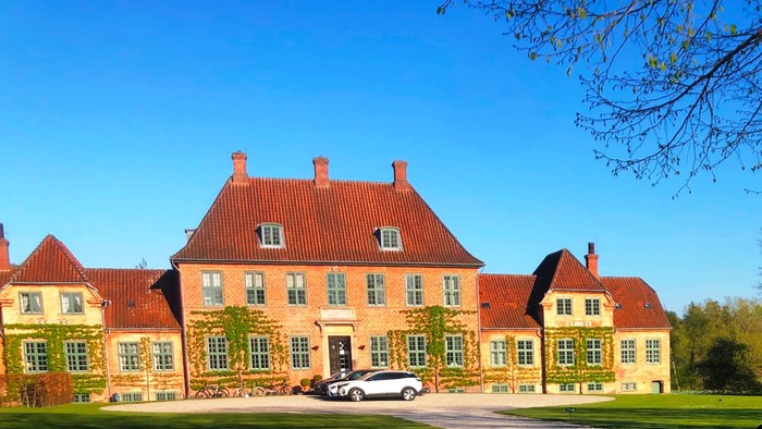 Elegant murstenspalæ med røde teglstenstag, frodig grønne omgivelser og en parkeret bil på en rummelig indkørsel.