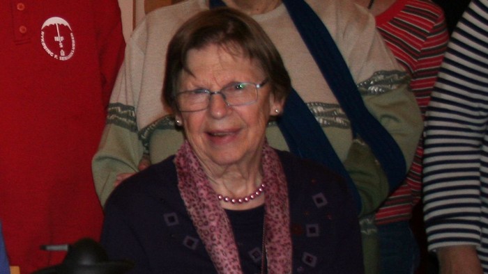 En glad ældre kvinde med briller og tørklæde omgivet af en gruppe mennesker til en begivenhed.