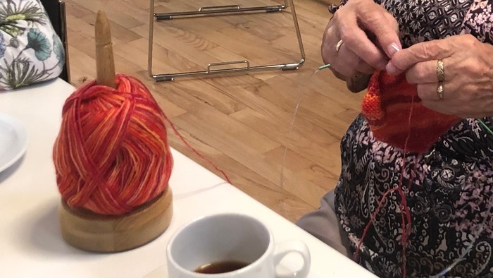 En gammel kvinde sitter og stråler med en rød tråde, mens andre folk sidder og snakker i baggrunden.