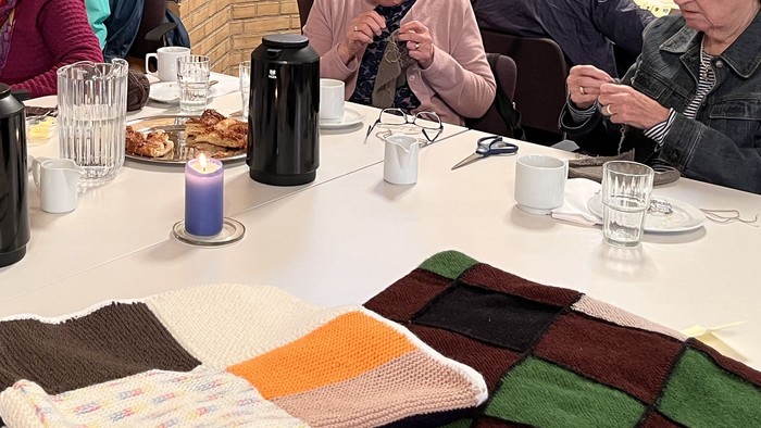 En gruppe ældre personer sidder ved et bord med te, kager og strikkearbejde.