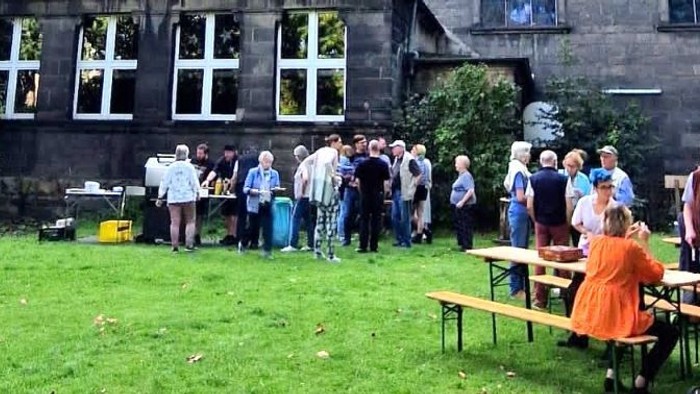 Gruppe von Menschen, die sich im Freien in der Nähe eines Gebäudes versammelt und eine gesellige Veranstaltung mit Picknicktischen und Grünflächen genießt.