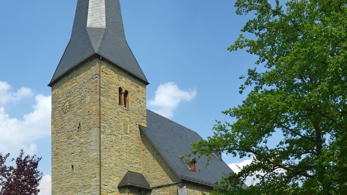 Auf dem Bild sieht man die Evangelische Kirche in Bad Sassendorf. Sankt Simon und Judas Thaddäus. Das Bild ist im Sommer aufgenommen worden. Die Sonne scheint, der Himmel ist blau und im Vordergrund sieht man grünen Rasen, Büsche und die große Eiche.