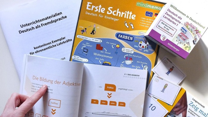 Eine Hand hält ein Buch mit dem Titel "Ersie Schrite", umgeben von verschiedenen Lernmaterialien und Karten.