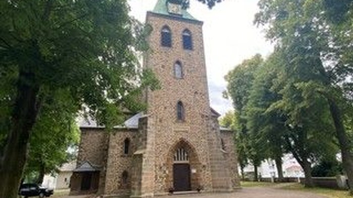 Das Bild zeigt eine steinerne Kirche mit hohem Turm und grünem Dach, umgeben von Bäumen und einem Parkplatz.