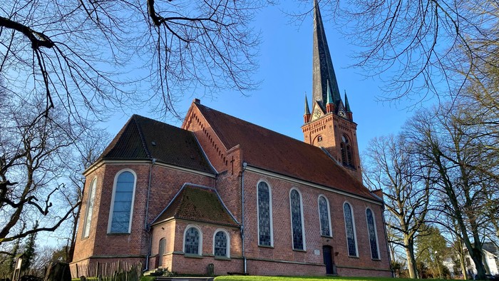 Kirchengebäude aus Ziegeln mit hohem Turm und spitzen Fenstern, umgeben von Bäumen und grünem Gras