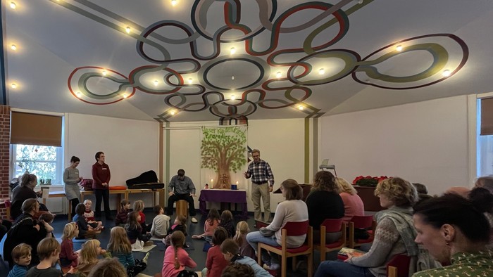 Eine Gruppe von Menschen sitzt in einem Raum mit kreisförmigen Deckenleuchten und hört einem Vortragenden zu.