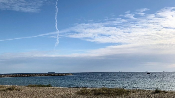 En smal røgspor fra et fly over en strand med lav vandstand og klart himmel.