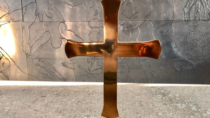 Guldfarvet kors på granitplade foran mørk mur med skitser.