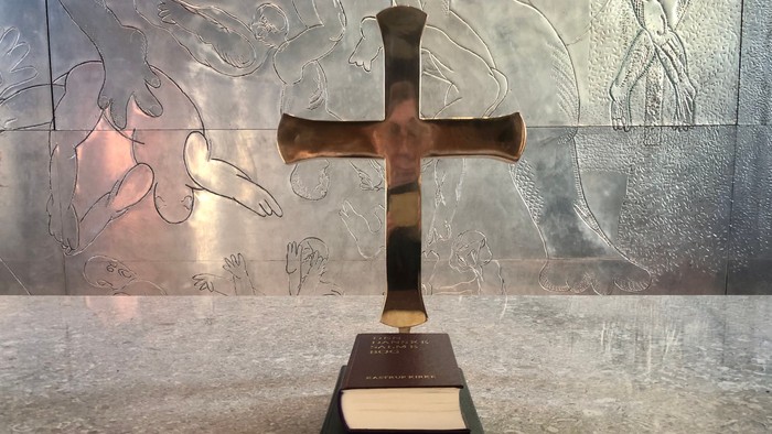 En stor kors står oven på en bundet bog på en poleret overflade med indgraverede håndaftryk i baggrunden.