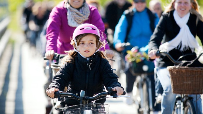 En gruppe cykelende på en bro, inklusive en lille pige med rosa hjelm.
