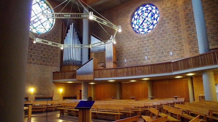 Moderne Kircheninnenausstattung mit hölzernen Kirchenbänken, einer Orgel und runden Buntglasfenstern mit warmem Licht.