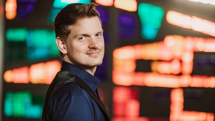 Ein Mann in dunklem Anzug vor farbigem Hintergrund.