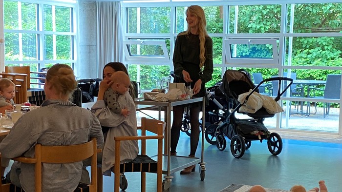 En gruppe kvinder sidder ved bord i et lys rum med store vinduer, mens deres børn spiller på gulvet.