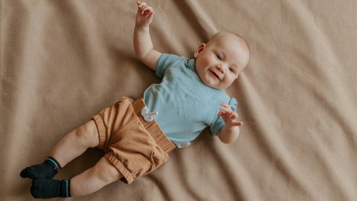 Et smilende baby i en lyseblå trøje og beige bukser ligger glad på en blød overflade.