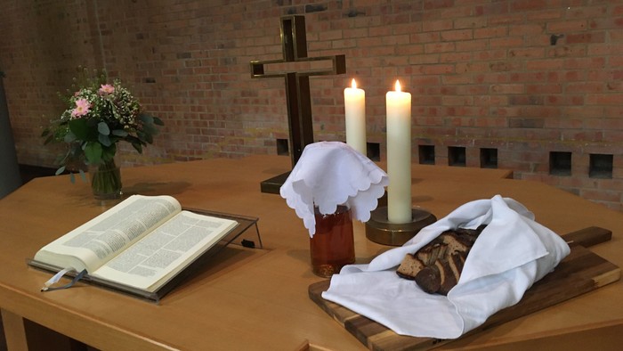 Religiöser Altaraufbau mit Kerzen, einem Kreuz, einem Buch, Blumen und einem bedeckten Laib Brot auf einem Holzbrett.
