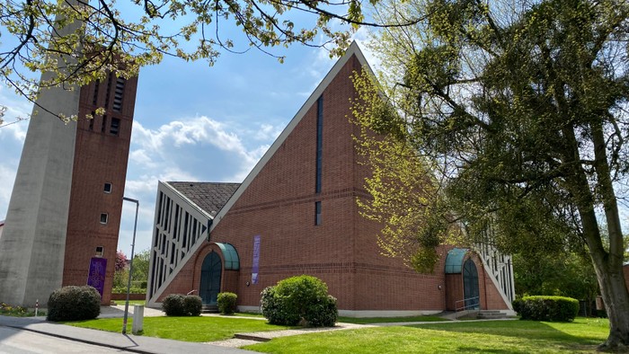 Moderne Kirche mit dreieckigem Design und hohem Turm, umgeben von Bäumen und grünem Rasen.