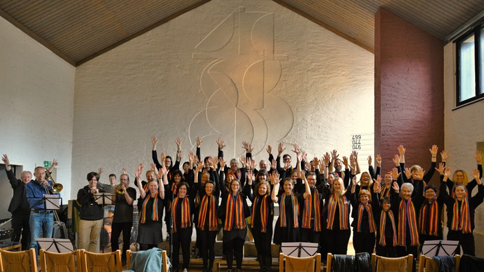 Ein großer Chor singt in einer Kirche, alle Mitglieder tragen orange-schwarze Outfits und heben die Arme, während ein Mann Notenblätter hält.