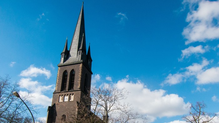 Ein hoher Kirchturm ragt vor einem klaren blauen Himmel mit einigen Wolken auf.