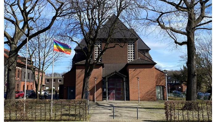 Bunte Flagge vor Backsteinkirche mit spitzem Dach und parkähnlichem Gelände