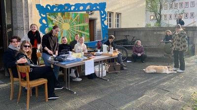 Gruppe versammelt sich im Freien um einen Tisch mit einem Hund und diskutiert oder organisiert etwas vor einem farbenfrohen Wandgemälde.