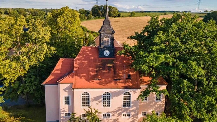 Kirche mit hohem Turm und Uhr, umgeben von Bäumen und Feldern