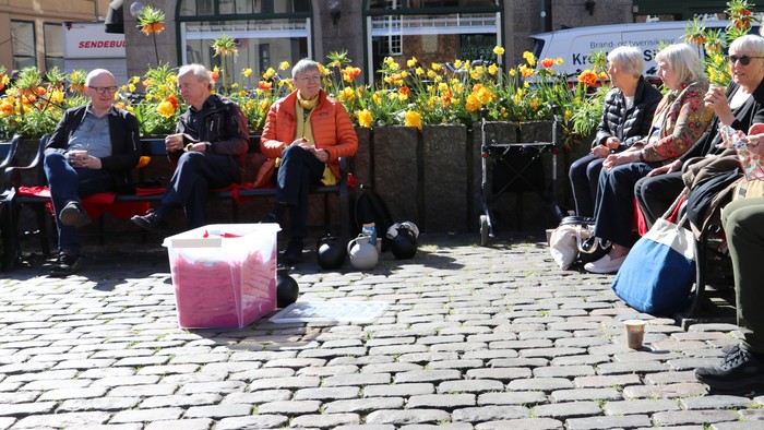 En gruppe pensionister sidder på bænke i en bygate med blomster og en rosa kasse foran dem