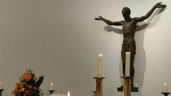 Eine feierliche Altarszene mit einer Kruzifix-Statue, brennenden Kerzen und Blumenarrangements auf einem bedeckten Tisch.