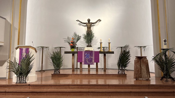 Ein ruhiger Kirchenaltar mit einem Kruzifix, Kerzen, Blumen und hölzernen Kirchenbänken unter einem großen Bogenfenster.
