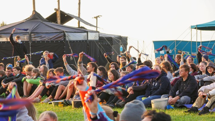 Menschenmenge, die auf dem Gras sitzt, einige halten Leuchtstäbe, im Hintergrund eine Bühne und Zelte.