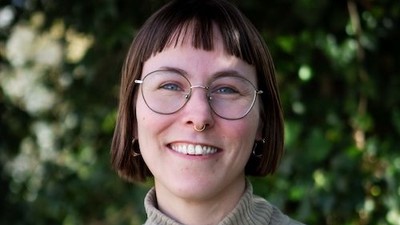 En glad person iført en farverig blomstermønstret sweater og briller, der smiler udendørs.