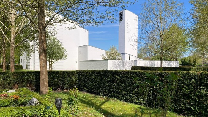 Et moderne, hvide kirke med høje tårne bag en grænse og en grøn have med træer og buske.