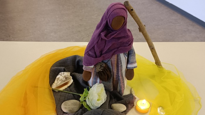 Eine Puppe in lila Kleidung sitzt auf einem Stoffhaufen, umgeben von Muscheln, einer Rose und Kerzen.