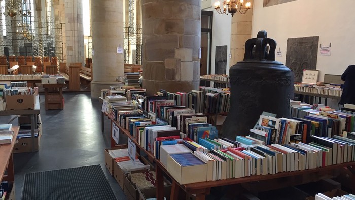 In der Bibliothek stehen viele Bücherregale mit Büchern, ein großer Raum mit hohen Fenstern und einem Kamin.