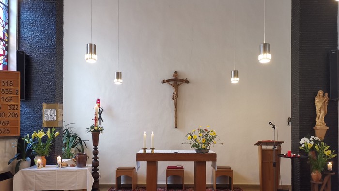 Ein ruhiger Kapelleninnenraum mit einem Altar, auf dem Kerzen, Blumen, ein Kruzifix und moderne Pendelleuchten zu sehen sind.