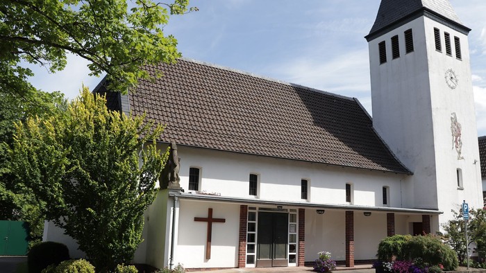 Kleine weiße Kirche mit schwarzem Dach und Uhrturm, umgeben von Bäumen und Büschen