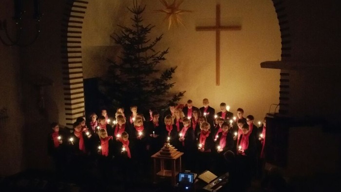 Der Gospelchor Talking People während eines Gottesdienstes. Die ca. 40 Sängerinnen und Sänger stehen im Altarraum der fast dunklen Adolf-Clarenbach-Kirche. Jeder Sänger hält eine Kerze in der Hand. Im Hintergrund sind ein Tannenbaum, Adventsstern und ein