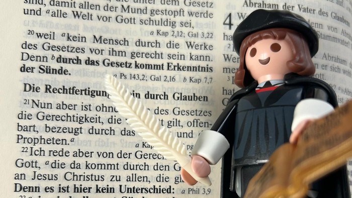 Eine Spielzeugfigur, die als Priester gekleidet ist, hält ein Buch vor einem deutschen Text.